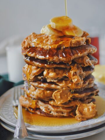 Peanut Butter Banana Pancakes stacked high, topped lots of crunchy peanut butter, banana slices, and maple syrup.