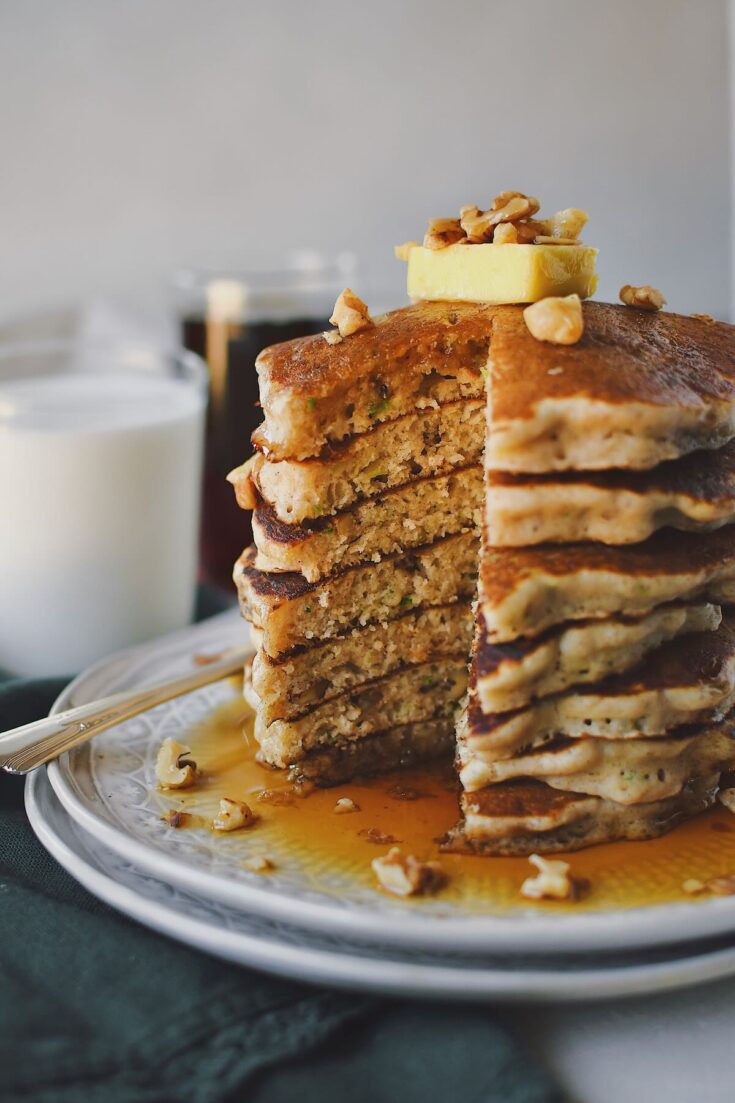 Zucchini Bread Pancakes stacked high and topped with a butter pat and extra walnuts. Sliced open showing the fluffy interior and zucchini shreds poking through.