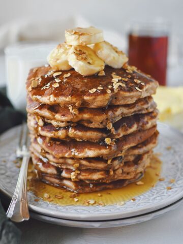 Banana Oat Pancakes stacked high, topped with banana slices and extra mini chocolate chips.