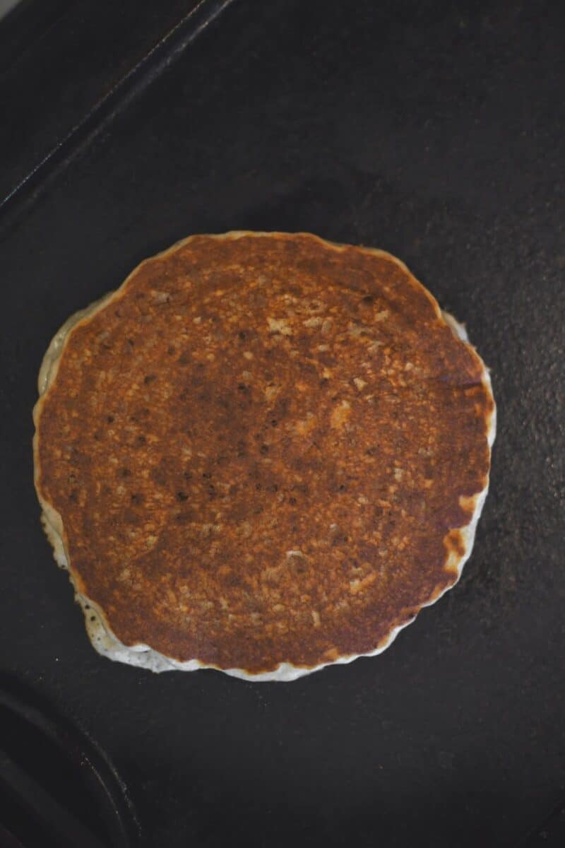 A pancakes cooking on a griddle, after flipping.