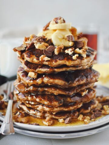 Banana Bread Pancakes stacked high, topped with sliced banana, walnuts, pecans, and chocolate chunks.