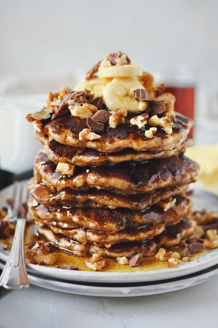 Banana Bread Pancakes stacked high, topped with sliced banana, walnuts, pecans, and chocolate chunks.