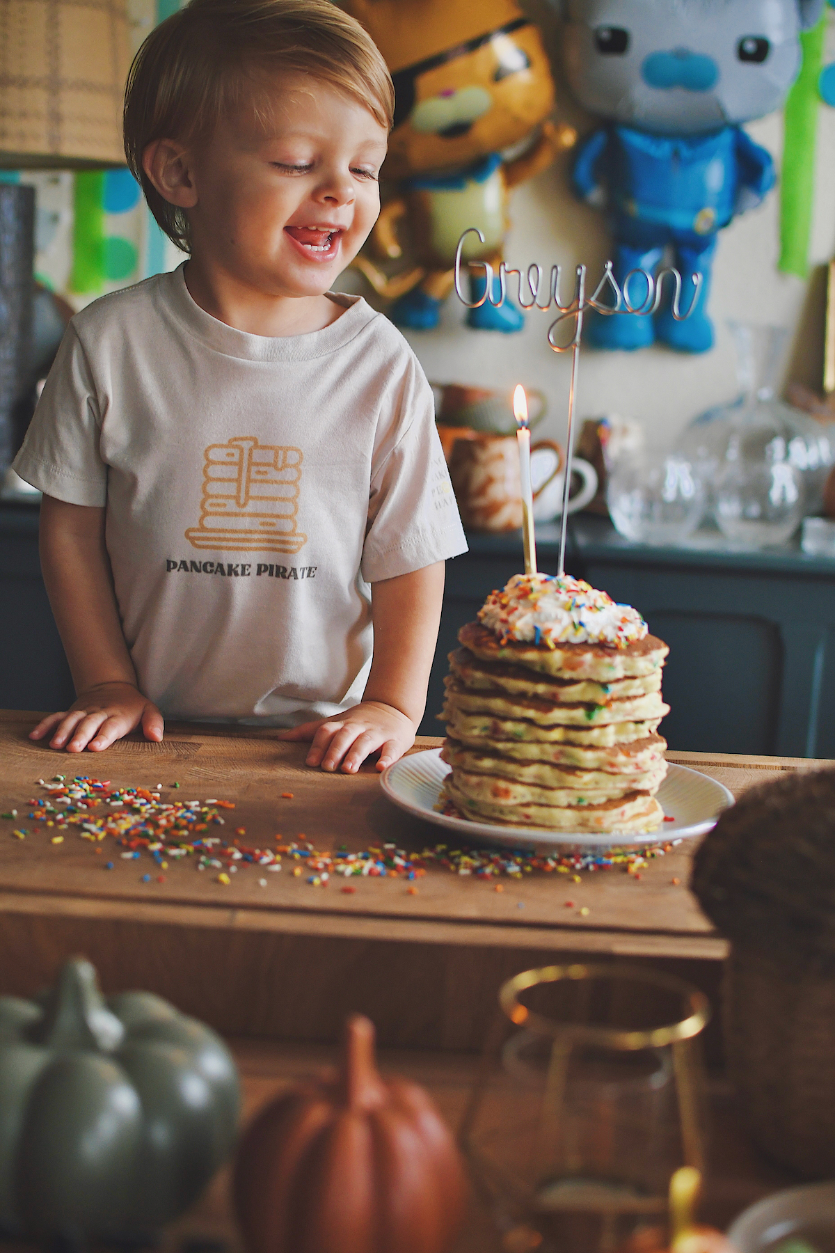 Birthday Cake Pancakes stacked up and topped with whipped cream and more sprinkles. A candle and a name pick in them. Greyson standing behind them.