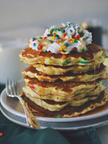 Funfetti Pancakes stacked up on a plate, topped with whipped cream, extra sprinkles, and dripping with maple syrup.