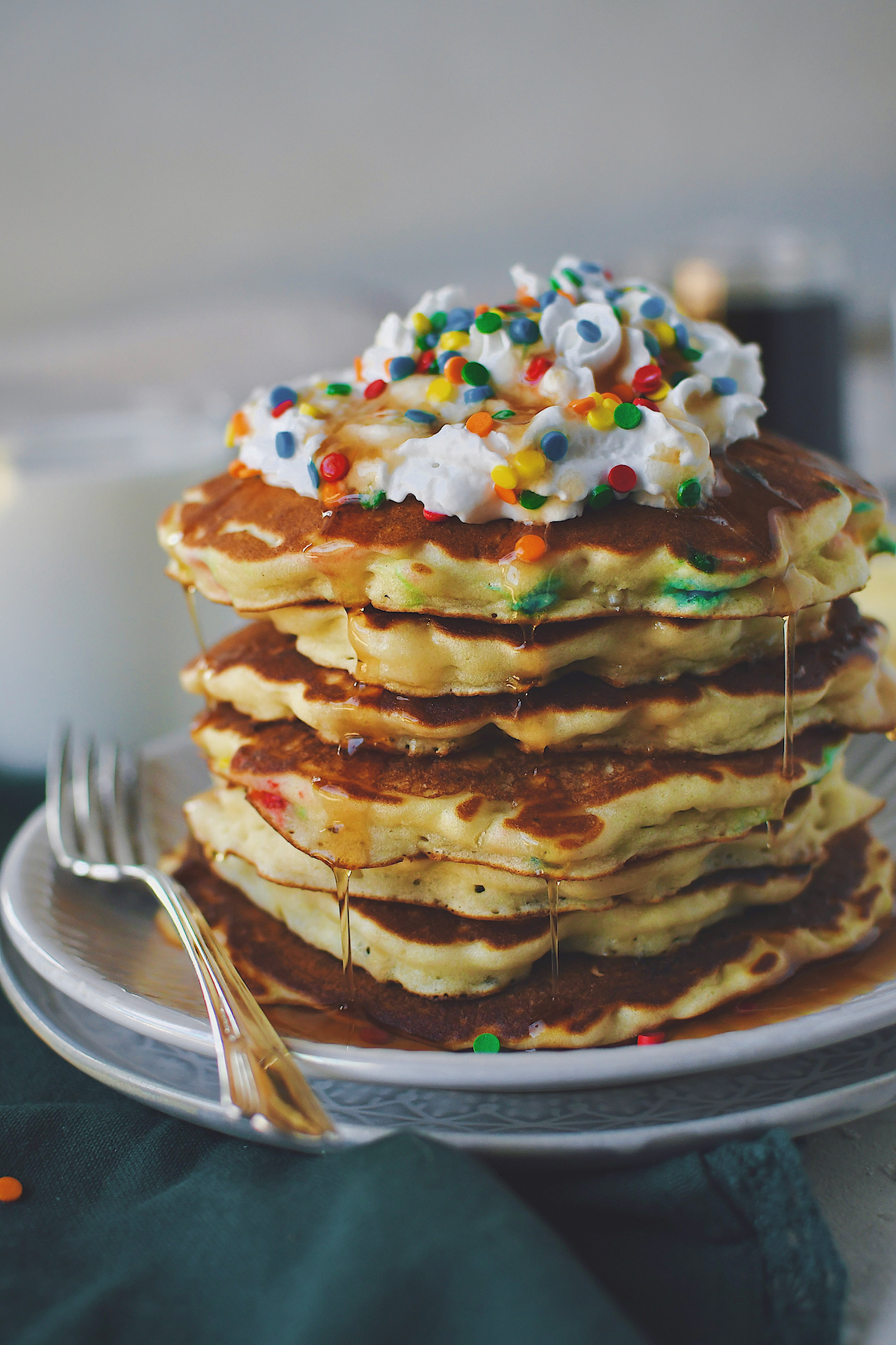 Funfetti Pancakes stacked up on a plate, topped with whipped cream, extra sprinkles, and dripping with maple syrup.