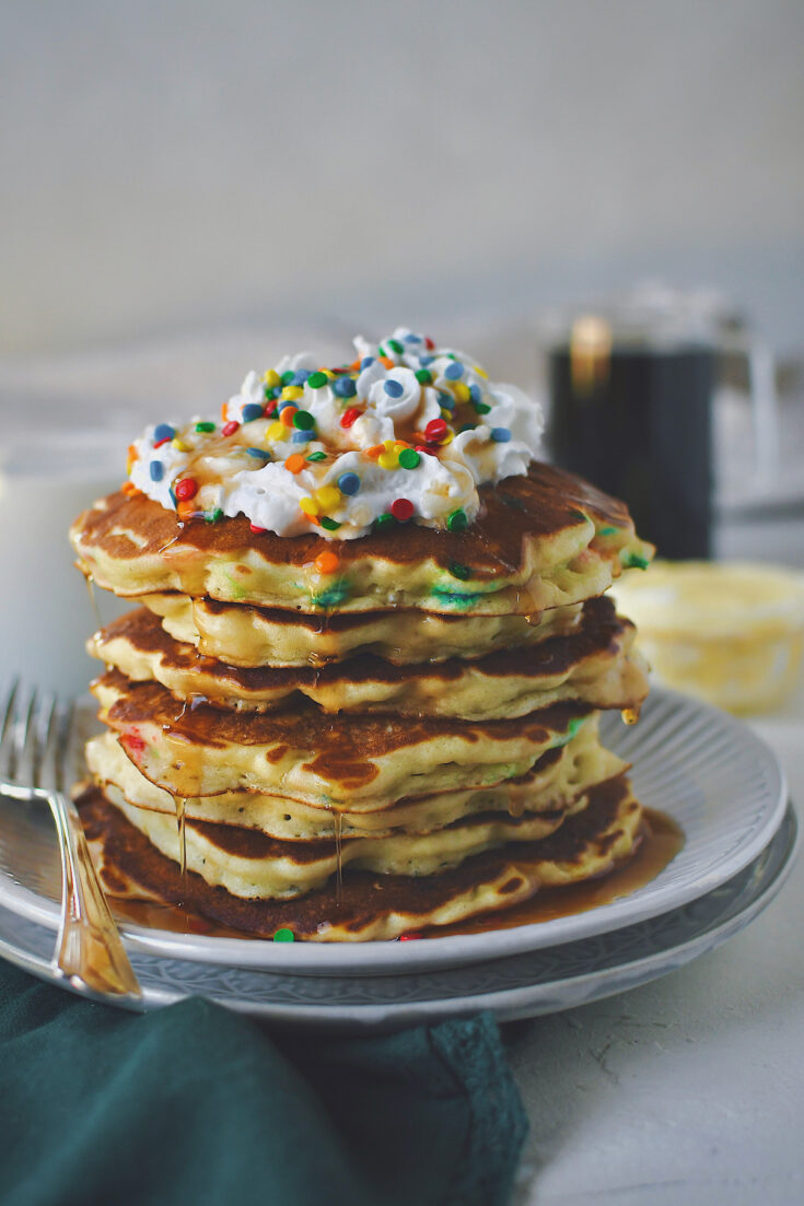 Funfetti Pancakes stacked up on a plate, topped with whipped cream, extra sprinkles, and dripping with maple syrup.