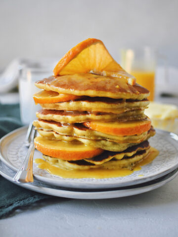 Orange Pancakes stacked high with some orange slices in them with a twist on top, and topped with orange maple syrup.