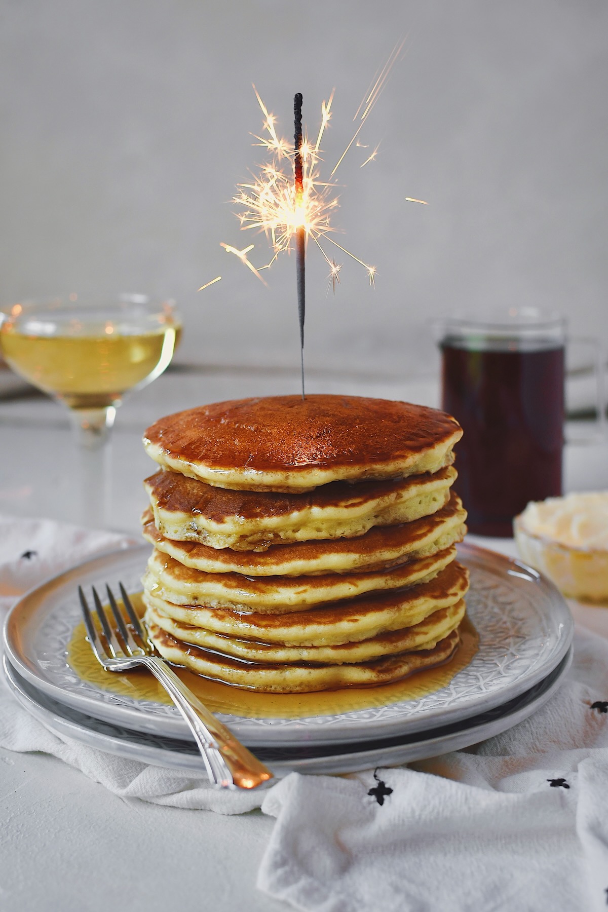 Champagne Pancakes stacked high, drizzled with maple syrup, served with a glass of champagne and a sparkler lit on top.