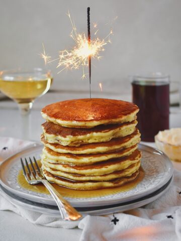 Champagne Pancakes stacked high, drizzled with maple syrup, served with a glass of champagne and a sparkler lit on top.