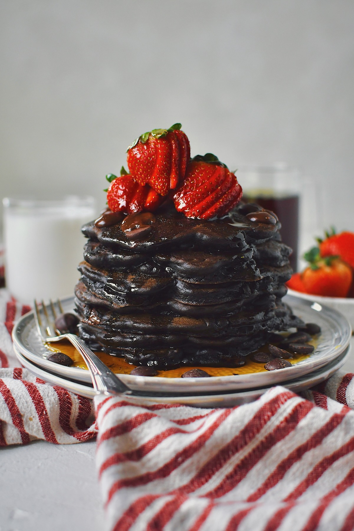 Chocolate Strawberry Pancakes stacked high, topped with sliced strawberries, extra chocolate chips, and a generous dose of maple syrup!