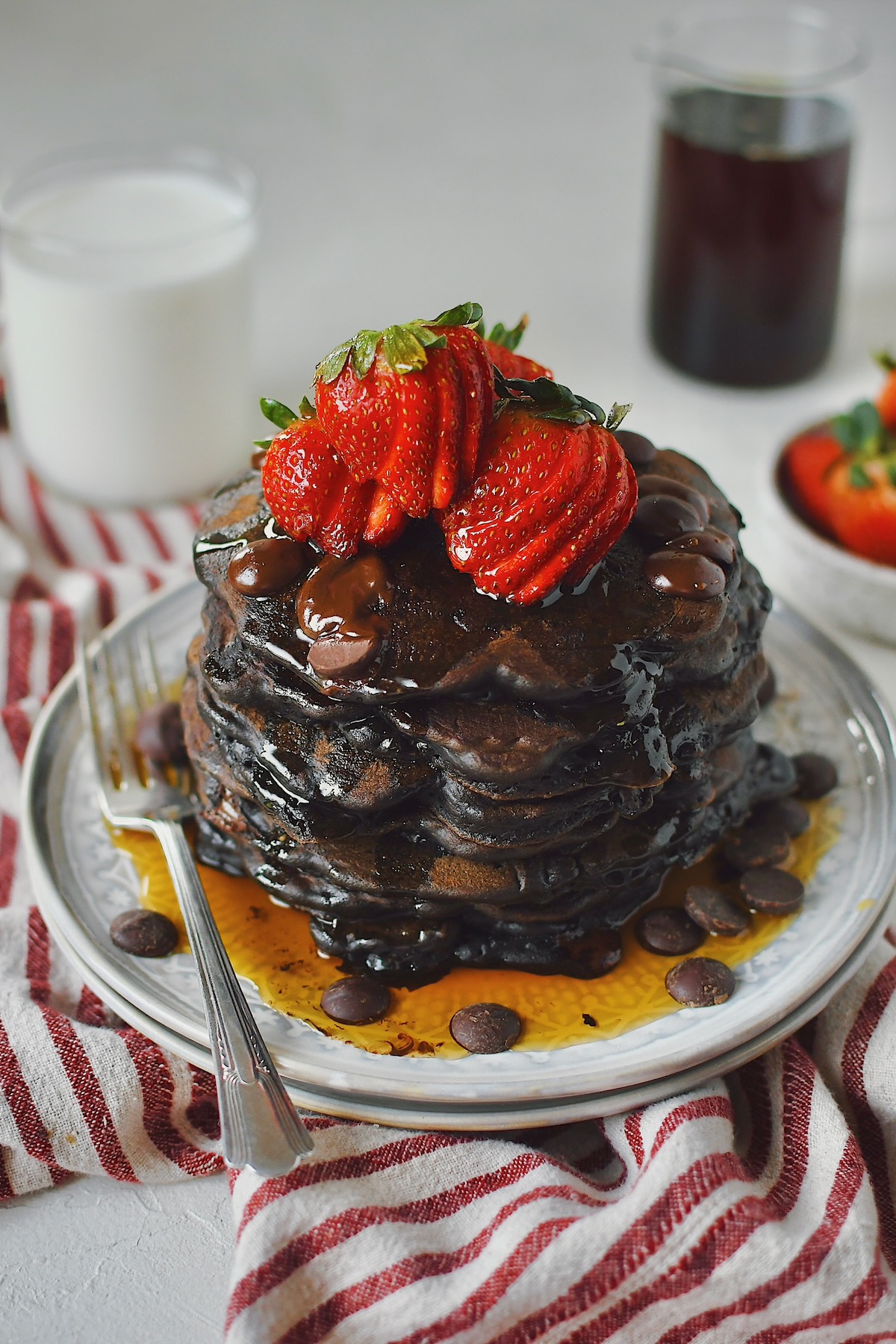 Chocolate Strawberry Pancakes stacked high, topped with sliced strawberries, extra chocolate chips, and a generous dose of maple syrup!