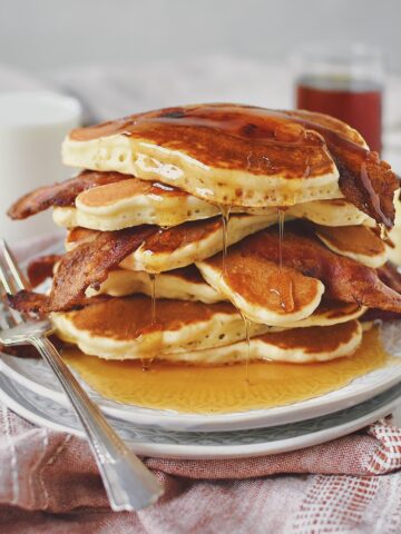 Bacon Pancakes stacked up on a plate with maple syrup DRIPPING off of them.